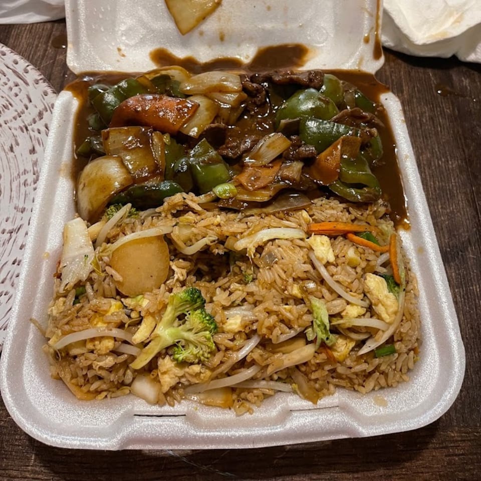 Chinese beef and peppers with fried rice in a white takeout container on a wooden table.