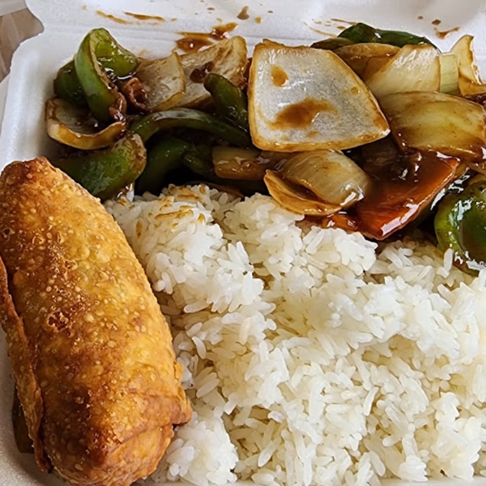 Chinese takeout meal with an egg roll, white rice, and pepper steak in a styrofoam container.